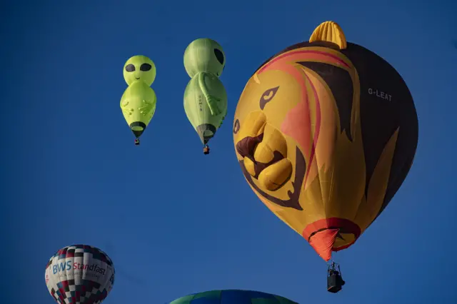An orange lion-shaped balloon hovers in the sky. Above it there are two green aliens.