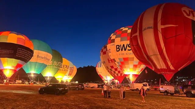 Under a dark blue sky several hot air balloons are lined up either side of the main arena at the Bristol Balloon Fiesta. They are being illuminated by their burners as part of the nightglow