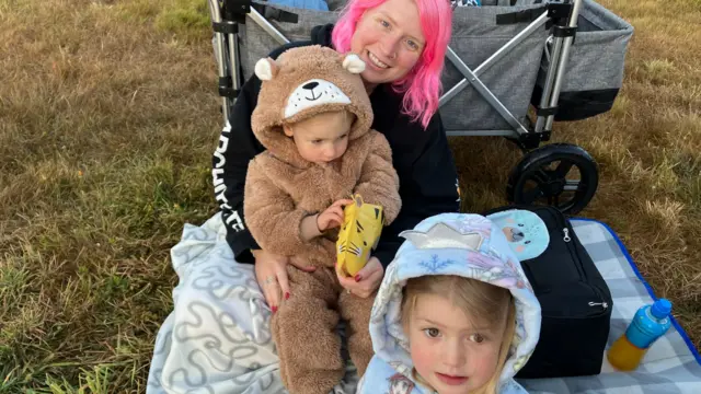 A woman with bright pink hair holds two small children on a picnic blanker. One of the children is in a large fluffy bear onesie, and the other is in a Frozen hoodie.