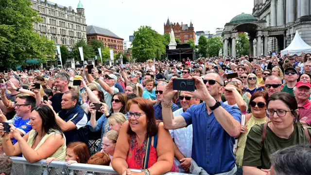 A large crowd of people. They are all looking in one direction. Several of them have their phones in their hands, recording/photographing what they are looking at. They are outdoors.