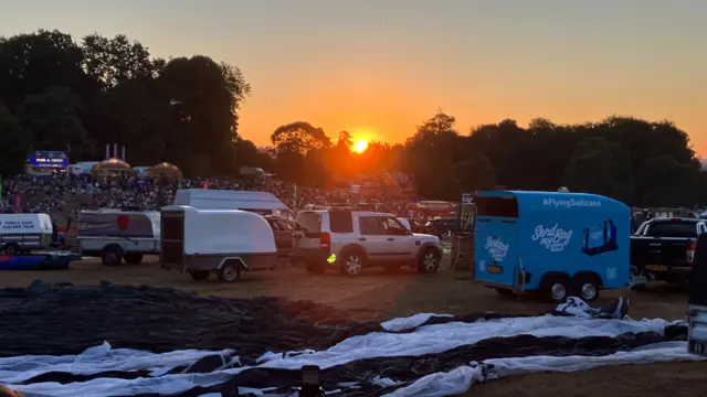 A bright orange sunrise peeks over the tree line at the fiesta site. There are lots of 4x4 trucks and trailers, with a balloon laid out in the foreground ready to be inflated.