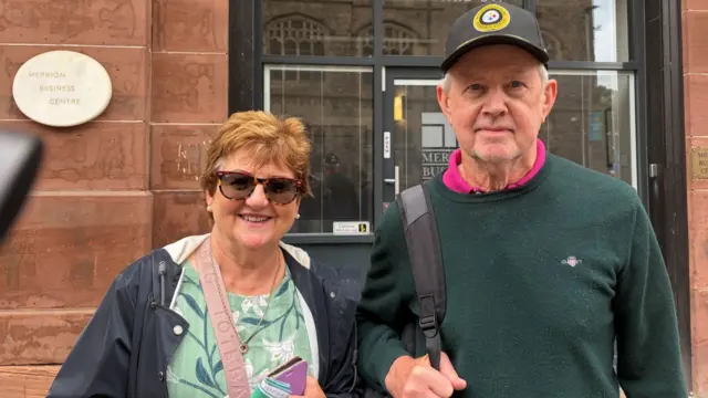 a man and woman looking at the camera. She is wearing a floral blouse, a rain jacket and sunglasses. He is wearing a pink polo shirt, a green jumper and a baseball cap.