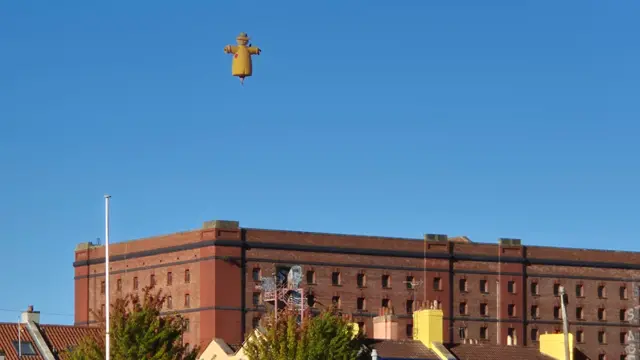 A yellow scarecrow balloon is flying above a reddish brown industrial building