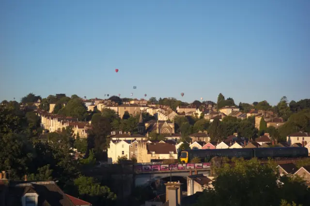 Balloons in the distance over houses. A train is running over a bridge in the foreground.