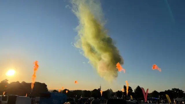 A large cloud of green smoke bursts into the blue sky. Orange flames can be seen reaching into the sky too, from the baskets.