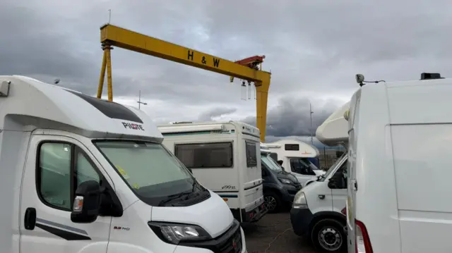 camper vans and motorhomes parked closely together, with a big yellow crane (labelled H&W) in the background