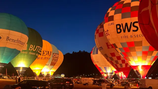 Several hot air balloons are lined up either side of the main arena at the Bristol Balloon Fiesta. They are being illuminated by their burners as part of the nightglow