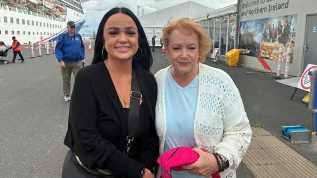 A woman wearing all balck with dark ahir and a woman wearing a blue top and white cardigan with red hair