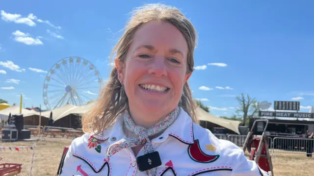 Dakota smiles at the camera with the fiesta grounds behind her. She has blonde hair and wears a western-style white shirt and a neckerchief.