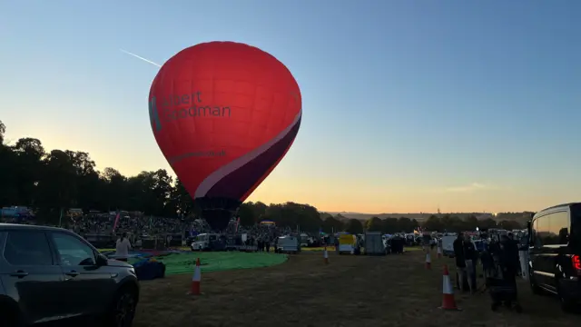 A red, black and white balloon inflated against a clear dawn sky. Crowds can be seen in the background.