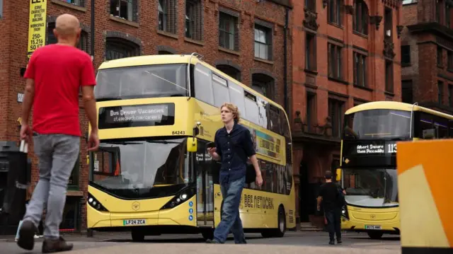 Two yellow buses can be seen in Manchester. Several people are walking past