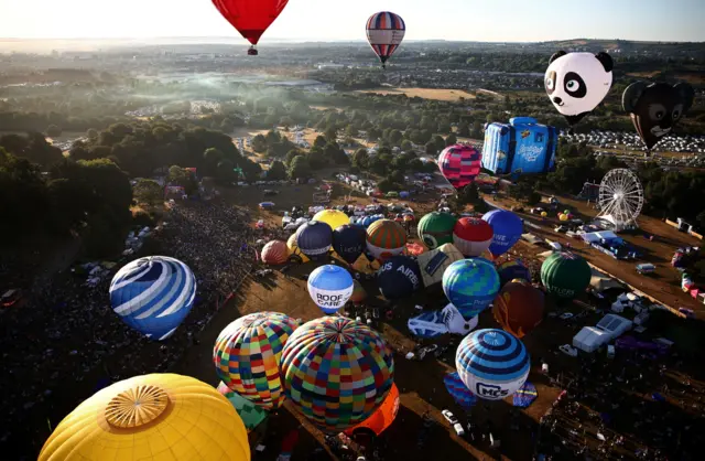 An aerial shot of the Bristol Balloon Fiesta site showing a dawn mass ascent. Dozens of colourful balloons are either inflating on the ground or already in flight