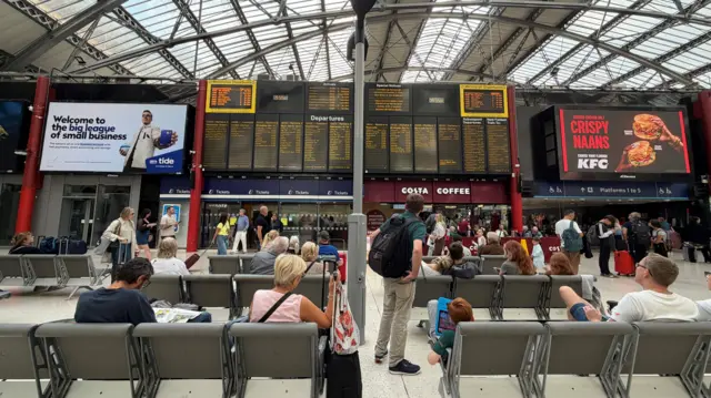 A departures board can be seen at Liverpool Lime Street. Several rows of chairs are in front of it and around a dozen people are looking at the train times