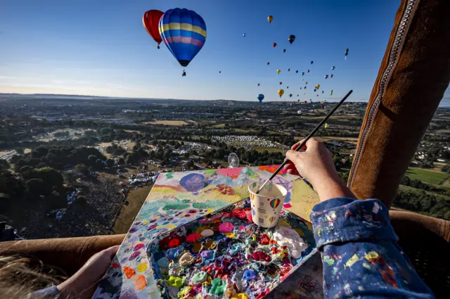 A colourful tray of paint rests on a canvas which is being worked on by an artist. The picture is taken from her perspective, and shows the landscape and balloons above it.