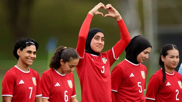 Five female football players are pictured in red shirts. The women in the centre holds her hands above her head in a heart gesture.