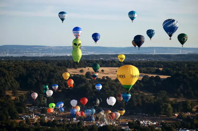 An aerial picture of a mass ascent at the Bristol Balloon Fiesta with dozens of colourful balloons in the air. The picture is taken looking towards the Seven Estuary and both river bridges are visible along with land on the Welsh side of the river