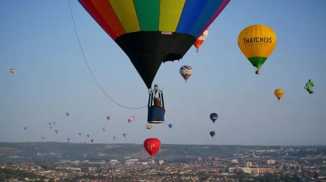 Dozens of balloons float in a bright blue sky across the city. There is one that can be seen up close, with a man operating the balloon.