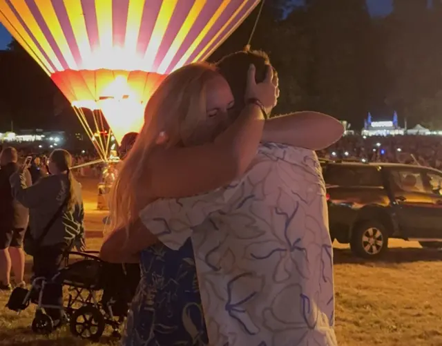 A man and a woman embrace in front of two hot air balloons at night