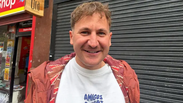 a man in a tee shirt and jacket smiling at the camera. A restaurant's closed shutters and a sign reading Orá are visible behind him