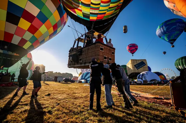 A group of people gather underneath the basket of a colourful hot air balloon as it takes off from Ashton Court