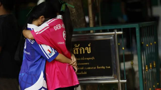Two children in brightly coloured sports tops embrace in front of a sign wirtten in Thai