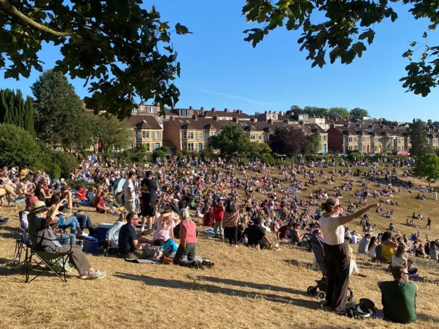 A large crowd of people gathered in a park on a sunny evening. The ground is light brown after weeks of heat and little rain