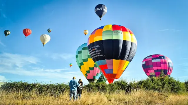 Multi-coloured balloons land in a field behind a hedgerow. There is a couple watching them come in to land.