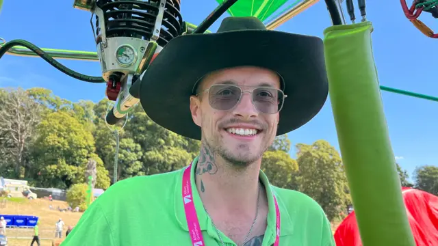 A man in a wide brimmed hat. sunglasses and a bright green t-shirt smiles from the basket of a hot air balloon which is tethered low to the ground.