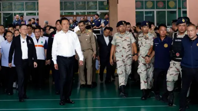 A large group of officials including some in military fatigues are shown arriving outside a school building - the prime minister walks at the front and is wearing dark trousers and a white shirt, with a somber look on his face.