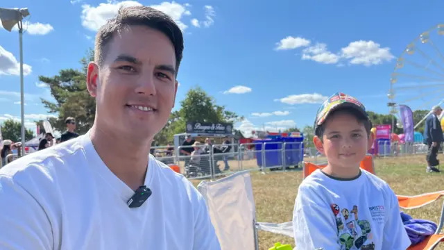 Carl, closest to the camera, smiles. He has short dark hair and wears a white t-shirt. Eli wears a white t-shirt with cartoons on it, and a multi-coloured cap.