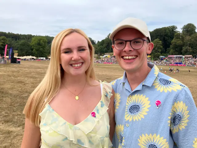 A young couple smile at the camera. She has long blonde hair and he is wearing a cap and glasses