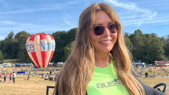 Carol Vorderman smiles in front of the balloon field, with a white and red striped balloon in the background. She has long, light brown hair, and wears dark glasses, a bright green t-shirt and a black cardigan.