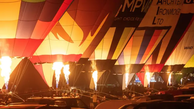 Seven hot air balloons tethered on the ground fire their burners as part of the nightglow at the Bristol Balloon Fiesta. The flames are bright orange