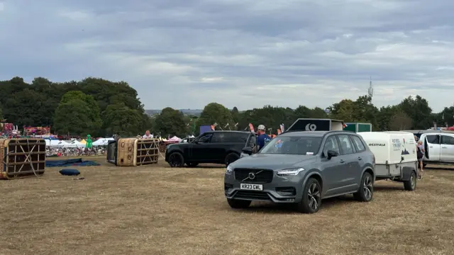 A grey Volvo in a field with a trailer on the back. There are several hot air balloon baskets turned on their side near it.