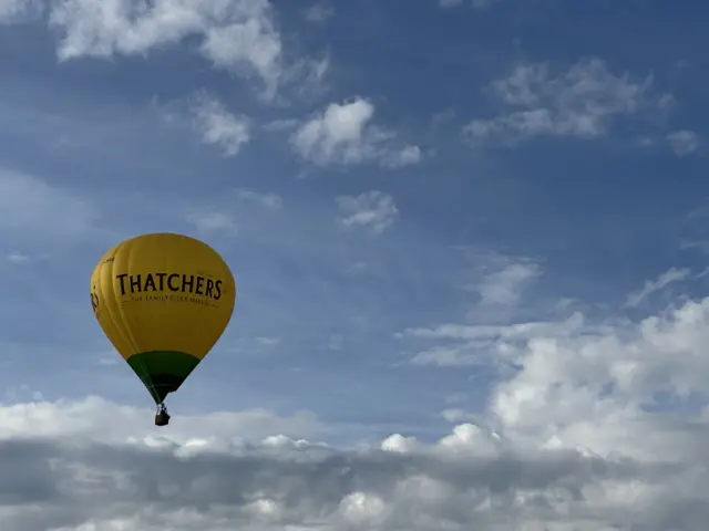 A yellow and green Thatchers cider balloon rises into a partly cloudy blue sky