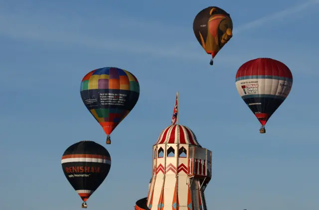 Four hot air balloons - one of which is in the shape of a lion - hover above a white and red helter skelter at a fairground