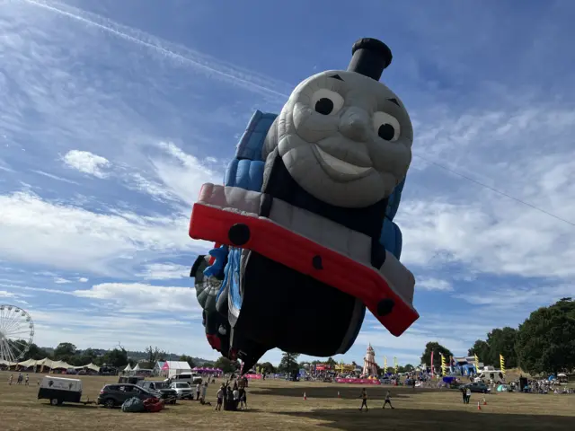 A huge blue and red Thomas the Tank Engine balloon inflates at the main field of the fiesta.
