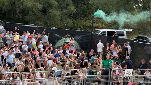 A crowd of people cheer and raise their arms as they see green smoke emerging from a pipe