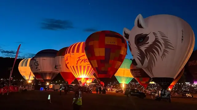 Hot air balloons - including one shaped like a wolf - stand in a line as they glow at night