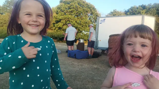 Two young girls smile and point at badges on their tops excitedly. Behind them a ballooning crew can be seen loading equipment into a trailer.