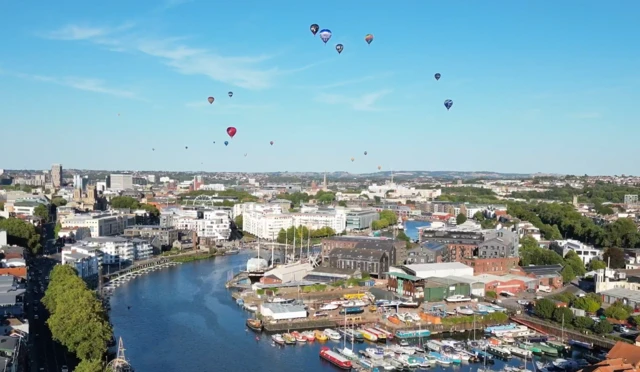 Hot air balloons hover in the distance above the harbourside. Bristol's skyline and a harbour with boats in it can be seen.