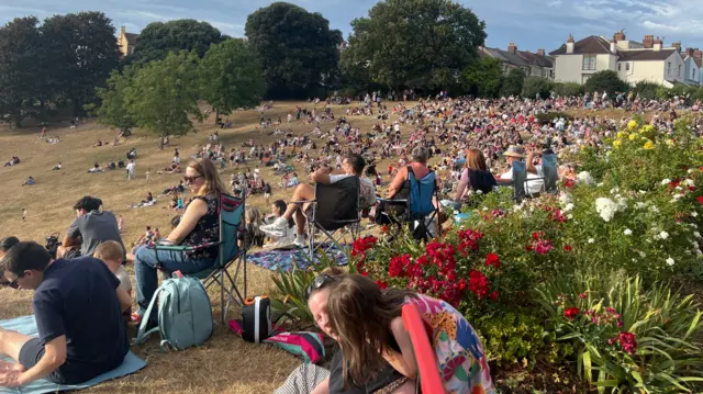 A large crowd sat down on a slope in a rather dry and crispy looking park in a residential area on a sunny evening.