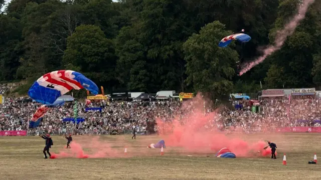 A group of RAF personnel landing after parachuting down from a plane. There is red smoke trailing behind them.