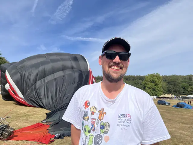 Gerard smiles widely at the camera, wearing a Bristol Balloon Fiesta top, dark glasses and a hat. The black and red Orient Express train is deflating behind him.