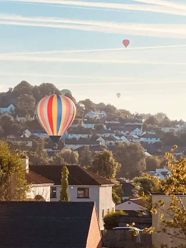 Three multi-coloured balloons pass over houses and trees.