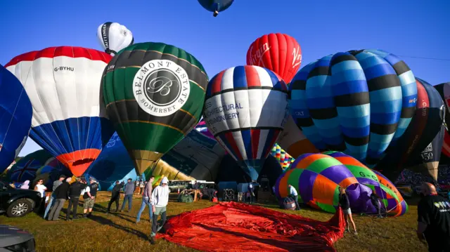 Several colourful hot air balloons are being inflated ahead of being launched on a sunny day at the Bristol Balloon Fiesta