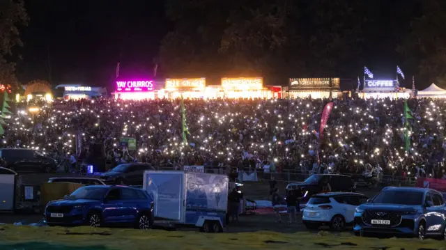 A large crowd is creating hundreds of individual points of light by holding up their mobile phones. In front of them are parked multiple large vehicles with hot air balloon boxes attached to them
