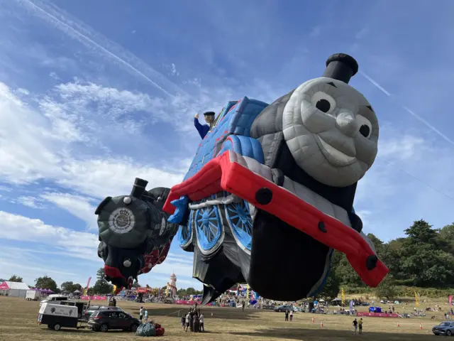 A huge blue and red Thomas the Tank Engine balloon inflates at the main field of the fiesta.