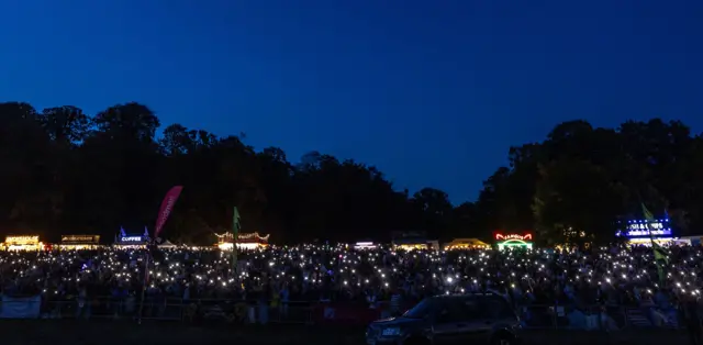 A large crowd holds up their phones with camera lights on at the Bristol Balloon Fiesta