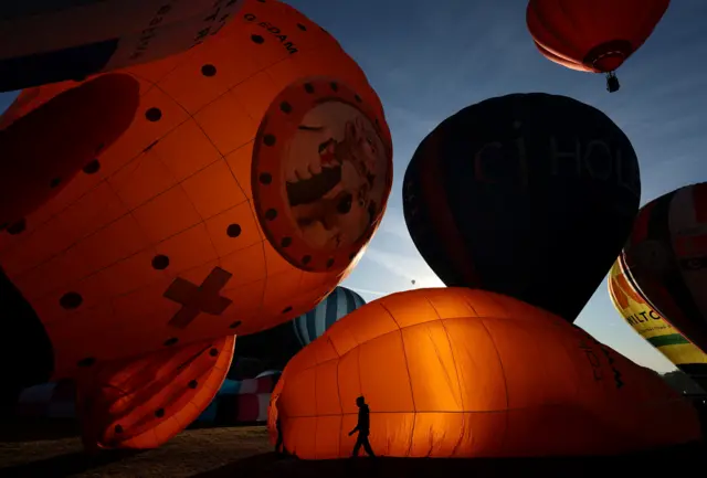 Lit-up hot air balloons are prepared for launch against an early morning blue sky. One of the balloons has a picture of Wallace and Gromit on it.